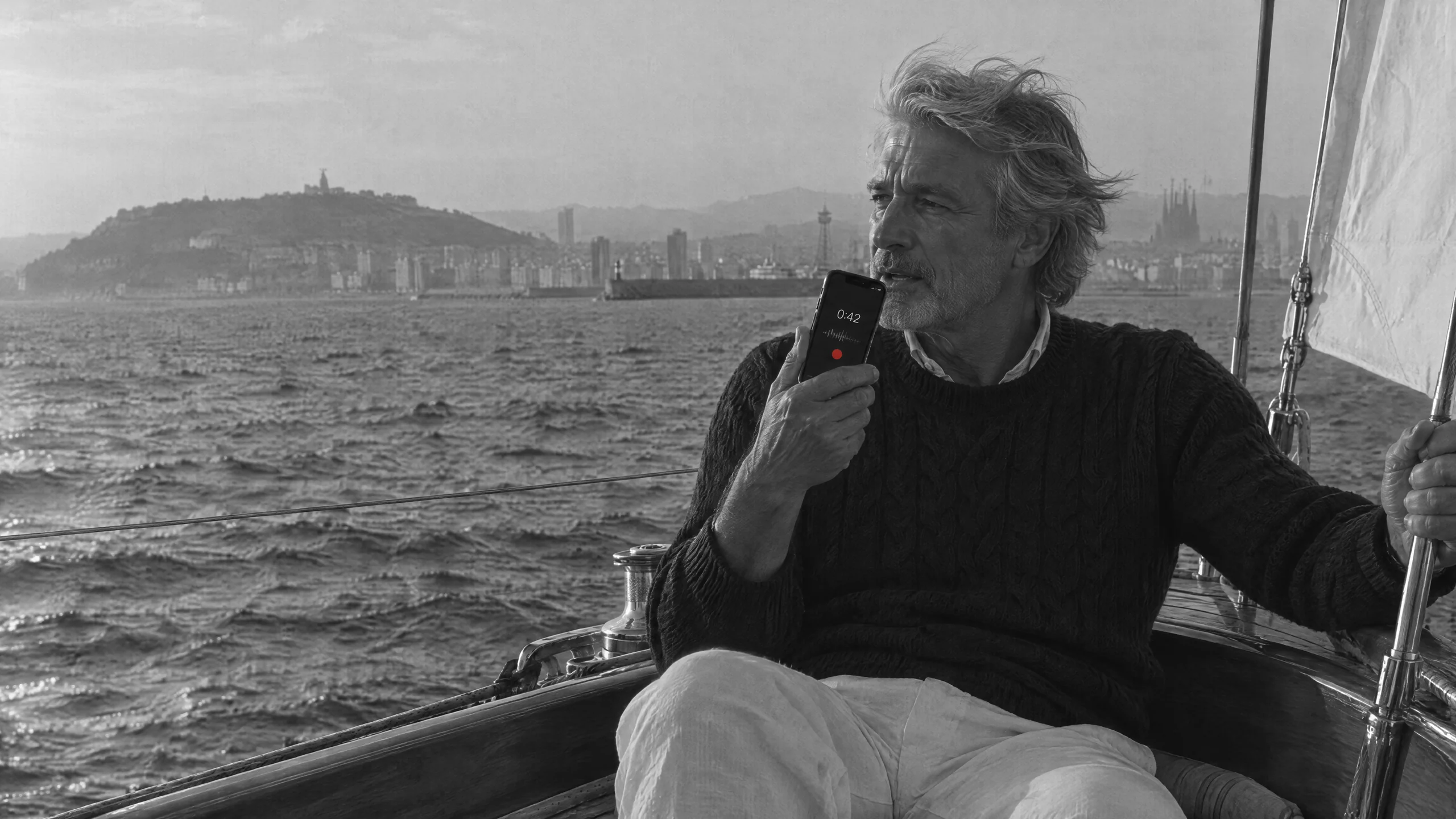 A silver-haired man on a teak sailboat off Barcelona at golden hour, speaking into his phone, Montjuïc and the Sagrada Família behind him