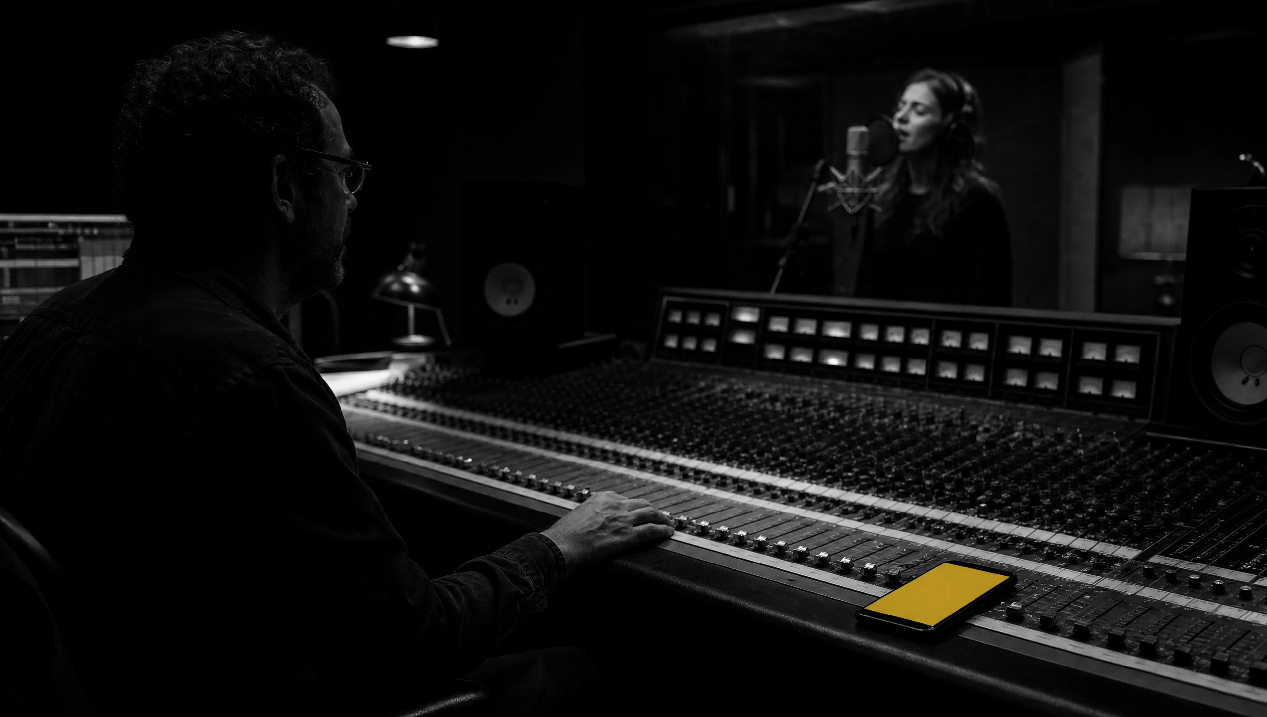 A producer at a mixing console watching a singer in the booth, his phone on the desk beside the faders